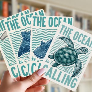 May include: A hand holds four rectangular cards with ocean-themed illustrations. Each card features a different marine animal: a whale, a stingray, a whale shark, and a sea turtle. The text "THE OCEAN is CALLING" is printed in teal.