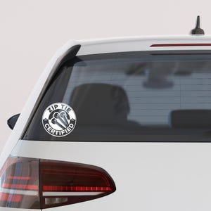 May include: White circular sticker on a car window. The sticker features the words "ZIP TIE CERTIFIED" in a circular design, with two zip ties in the center. The sticker is black and white.