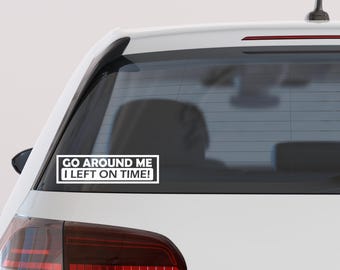 May include: White rectangular bumper sticker with black text that reads "GO AROUND ME I LEFT ON TIME!" The sticker is affixed to the rear window of a white car. The car's taillight is visible.