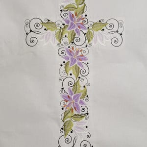 May include: Embroidered cross design featuring purple lilies, green leaves, and white flowers. The cross is outlined with black swirls and dots, set against a white background. A decorative religious item.