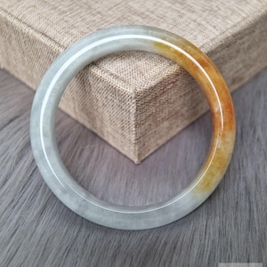 Op de afbeelding: Een gladde, ronde jade armband met een kleurverloop van bleek, doorschijnend grijsachtig wit naar een warm, levendig amberoranje. Het oppervlak van de steen is gepolijst en reflecterend, wat de natuurlijke minerale variaties benadrukt.