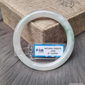 以下が含まれることがあります： 淡い緑色のジェダイトバングルブレスレット。ブレスレットは、背景に箱がある灰色のテクスチャード加工された表面に表示されます。「NATURAL JADEITE JADE」と「N°：345006」と書かれたタグが付いています。