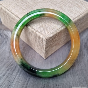May include: A smooth, circular bangle bracelet featuring a polished finish with a variegated color pattern of vibrant green, deep emerald, and warm amber-orange tones. The surface is translucent and reflective, showcasing natural color variations.
