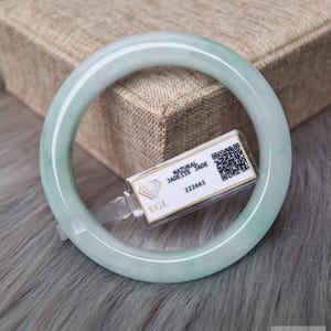 May include: A smooth, circular bangle bracelet crafted from pale green natural jadeite jade. A small white rectangular tag is attached to the bangle, featuring the text Natural Jadeite Jade, the number 222661, an EGL logo, and a QR code.