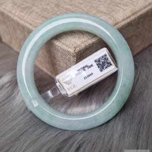 May include: A smooth, pale green jadeite bangle bracelet with a translucent appearance. A white rectangular tag is attached to the bangle, featuring the text Natural Jadeite, EGL, 222664, and a QR code for authentication purposes.