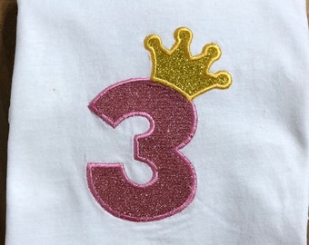 Peut inclure: T-shirt blanc avec un chiffre 3 rose avec des paillettes dorées et une couronne dorée à paillettes sur le dessus.