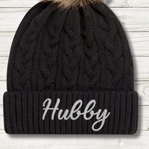 May include: Black cable knit beanie with a brown faux fur pom-pom. The word "Hubby" is embroidered in white script across the front. The hat is designed for cold weather.