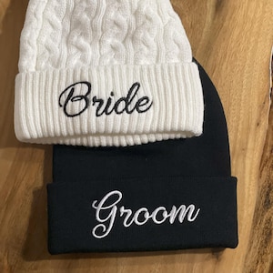 May include: A white cable knit beanie with a brown faux fur pom-pom and the word "Bride" embroidered in black script sits atop a black beanie with "Groom" embroidered in white script. The hats are likely for a wedding.