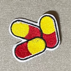 May include: Embroidered patch featuring three capsules. Each capsule has a red body and a yellow center, outlined in black and white. The patch is likely for clothing or accessories.