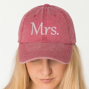 Peut inclure: Casquette de baseball rose poudré avec le mot "Mrs." brodé en blanc. La casquette a une visière incurvée et une conception classique à six panneaux. Le tissu a un aspect légèrement délavé et vintage.