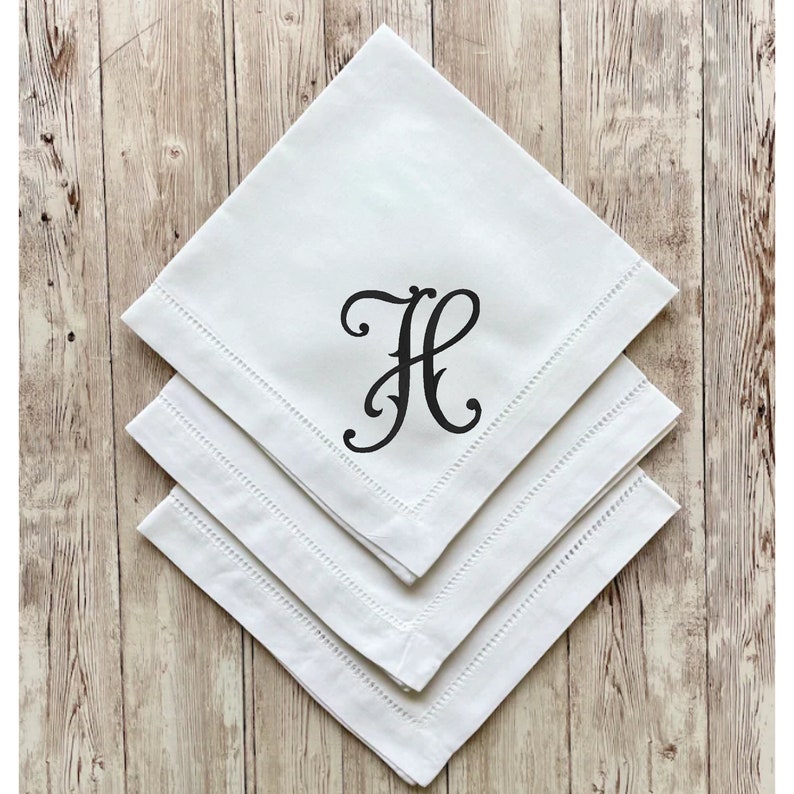 May include: Three white linen napkins with a decorative stitched edge are stacked on a wooden surface. Each napkin is folded into a square and features a black, stylized letter "H" in the center.
