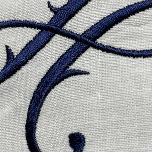 May include: Close-up of an embroidered navy blue letter "F" on a light gray linen fabric. The elegant cursive letter is intricately stitched, showcasing a detailed design. The fabric has a subtle texture.