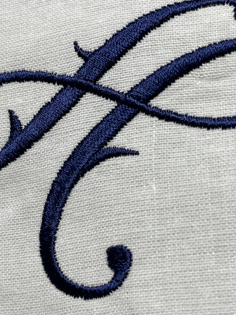 May include: Close-up of an embroidered navy blue letter "F" on a light gray linen fabric. The elegant cursive letter is intricately stitched, showcasing a detailed design. The fabric has a subtle texture.