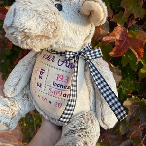 Puede incluir: Un peluche de alce en tonos beige y marrón claro, con una cinta a cuadros en blanco y negro. El alce tiene detalles bordados y un parche con el nombre "Carter Ann", peso, hora y 49 cm.