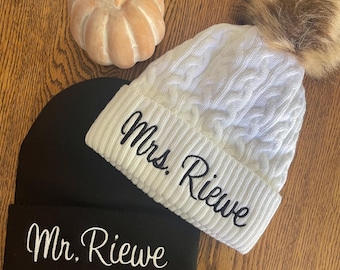 May include: A black beanie with "Mr. Riewe" embroidered in white script sits next to a white cable knit beanie with "Mrs. Riewe" embroidered in black script. A faux fur pom-pom tops the white hat. A small, light orange pumpkin decoration is also present.