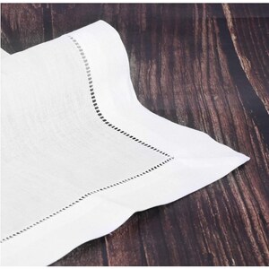 May include: A white linen napkin with a decorative stitched border. The napkin is folded, showing a corner with a detailed, perforated edge. The fabric has a subtle texture, and the background is a dark wood surface.