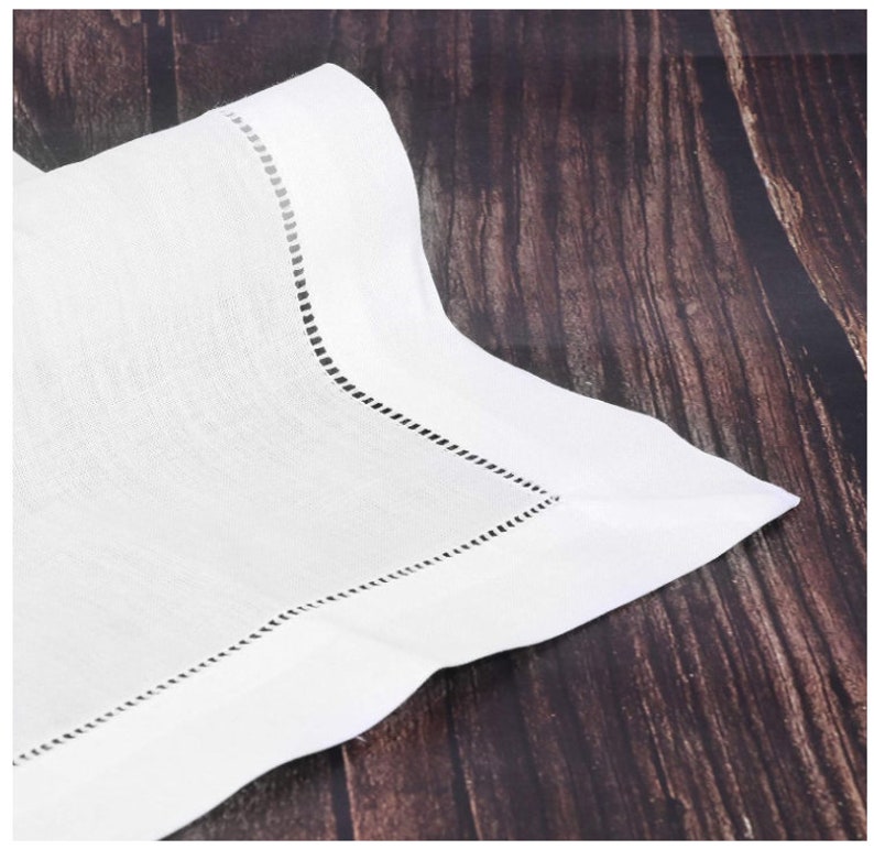 May include: A white linen napkin with a decorative stitched border. The napkin is folded, showing a corner with a detailed, perforated edge. The fabric has a subtle texture, and the background is a dark wood surface.