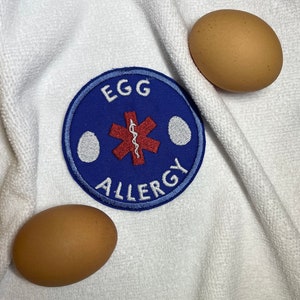 May include: A blue and white embroidered patch with the words "Egg Allergy" and a red medical symbol. Two brown eggs are on a white fabric background.