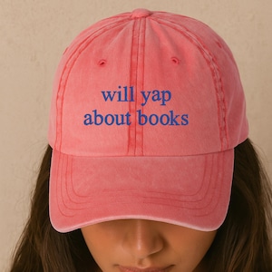 May include: A coral-colored baseball cap with the text "will yap about books" in blue. The cap has a curved brim and a classic six-panel design. The hat is made of a soft, washed fabric.