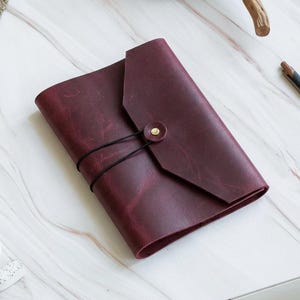 Puede incluir: Un diario de cuero color burdeos profundo con un diseño de cubierta envolvente, asegurado por un cordón elástico negro fino que se engancha en un pequeño botón de metal color latón. La superficie de cuero muestra texturas naturales.