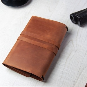 May include: A brown leather journal with a strap closure. The journal is closed and lying flat on a white surface.