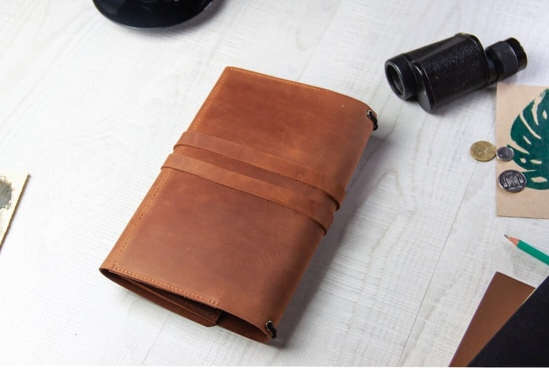 May include: A brown leather journal with a strap closure. The journal is closed and lying flat on a white surface.