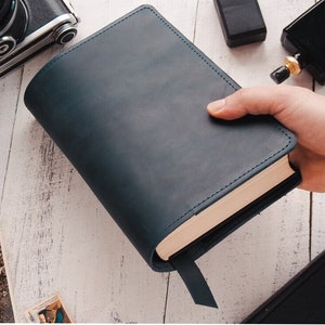 May include: A dark blue leather-bound book with a cream-colored spine and a gold-colored bookmark. The book is being held by a hand.