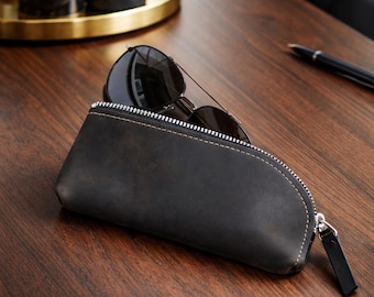 May include: A dark charcoal gray leather zippered case with a curved top edge and light-colored stitching. The case is partially open, revealing a pair of dark-tinted aviator sunglasses with thin metal frames resting inside the pouch.