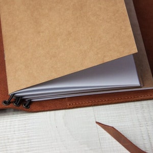 May include: A brown leather bound journal with a blank page open. The journal has a metal clasp and is closed with a brown leather strap.