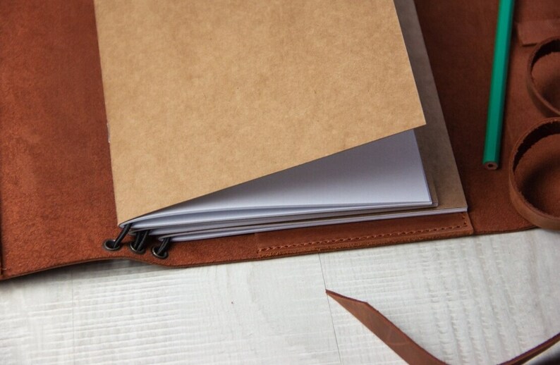 May include: A brown leather bound journal with a blank page open. The journal has a metal clasp and is closed with a brown leather strap.