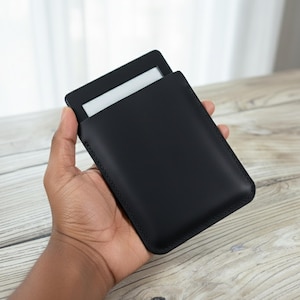 May include: A black leather e-reader case with a white screen visible. The case is held in a hand, highlighting its simple design. The case is designed to protect the e-reader. The case is made of leather.