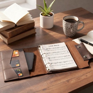 Può includere: Un raccoglitore in pelle marrone con uno smartphone e carte di credito nella tasca anteriore. Il raccoglitore è aperto per rivelare un raccoglitore con spartiti musicali con l'etichetta "Love Story". Una tazza di caffè, libri e una piccola pianta in vaso sono anche sulla superficie di legno.