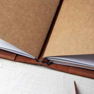 May include: A brown leather bound journal with blank pages. The journal is open and the pages are folded over. The journal is held together with metal rings.