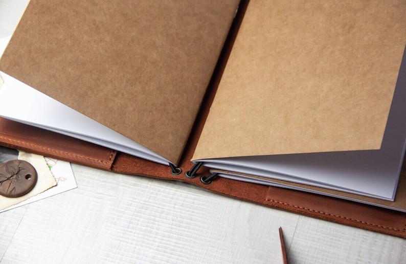 May include: A brown leather bound journal with blank pages. The journal is open and the pages are folded over. The journal is held together with metal rings.