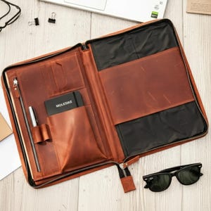 Puede incluir: Portafolio de cuero marrón abierto con cierre de cremallera, que contiene un cuaderno, un bolígrafo y ranuras para tarjetas. El portafolio está sobre una superficie de madera clara, con gafas de sol negras y material de oficina cerca. El cuaderno está etiquetado "MOLESKINE".