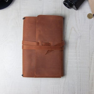 May include: A brown leather journal with a strap closure. The journal is closed and lying flat on a white wooden surface.