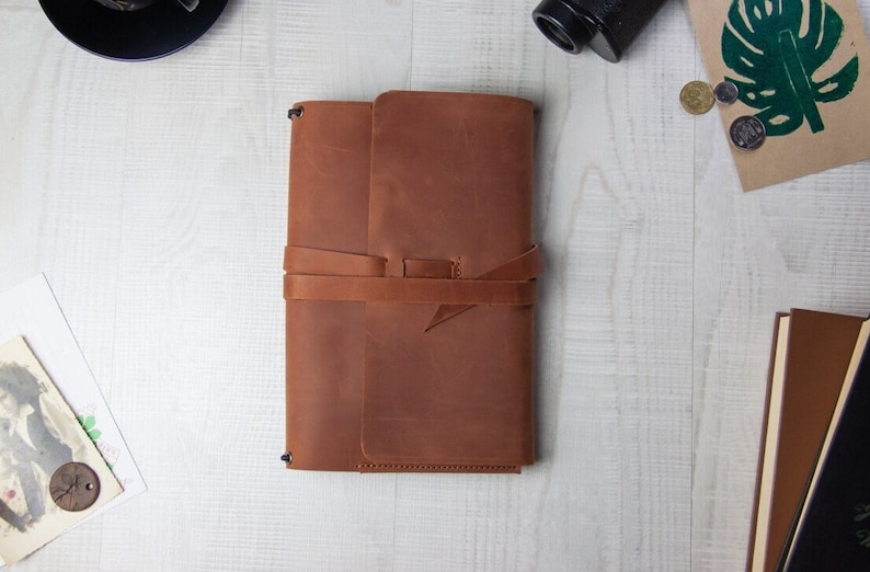 May include: A brown leather journal with a strap closure. The journal is closed and lying flat on a white wooden surface.