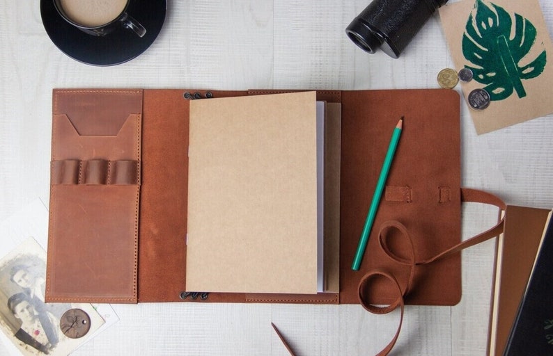 May include: A brown leather journal with a blank notebook inside. The journal has a pen holder and a strap closure. A green pencil is lying on the journal.