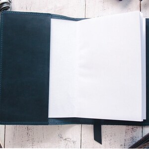 May include: A dark blue leather-bound journal with blank pages, open to reveal the white paper inside. A black pen is lying on a white surface next to the journal.