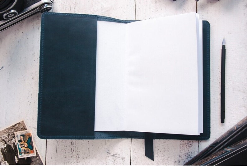 May include: A dark blue leather-bound journal with blank pages, open to reveal the white paper inside. A black pen is lying on a white surface next to the journal.