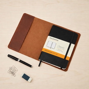 Op de afbeelding: Een bruine lederen notitieboekhoes met een zwart Moleskine Classic Collection gelinieerd notitieboek met elastiek, vergezeld van een zwarte balpen, een kleine rechthoekige gum en een stapeltje zilverkleurige metalen paperclips.