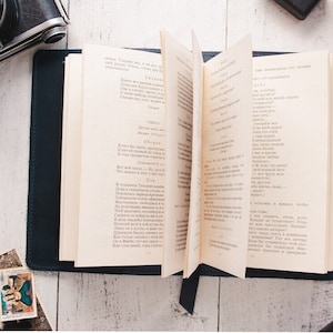 May include: A blue leather-bound journal with a bookmark, open to a page of text in a foreign language. The journal is on a white wooden surface with a vintage camera and a photograph.