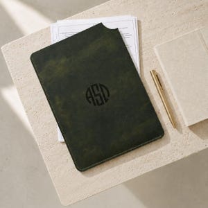 May include: A dark olive green leather document holder featuring a circular monogram with the letters ASD embossed in the center. The rectangular sleeve contains white paper documents and is positioned next to a gold metallic pen and a notebook.