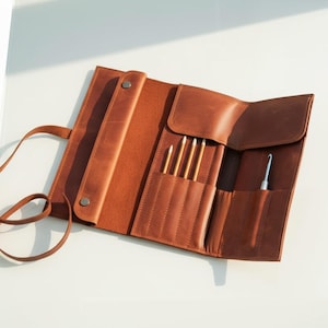 May include: Open, brown leather crochet hook and needle roll. The roll is partially unrolled, revealing pockets holding crochet hooks and needles. A strap is attached for securing the roll. The leather has a natural, textured appearance.