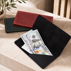 May include: A black leather checkbook cover is open, revealing a one hundred dollar bill tucked inside. Two additional checkbook covers, one in black and one in deep red, are positioned closed in the background, showcasing a matte finish.