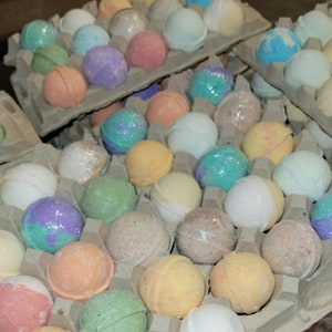 May include: Assortment of colorful bath bombs in various shades of pink, purple, blue, green, and yellow. The bath bombs are arranged in cardboard egg cartons. Some are wrapped in clear plastic.