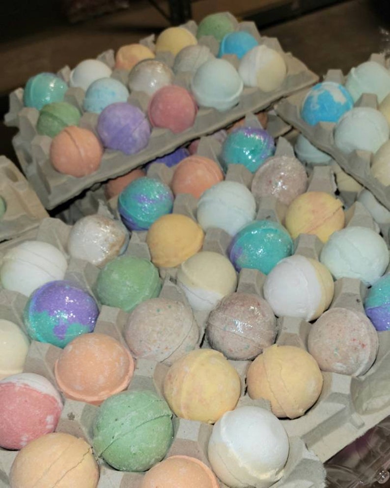 May include: Assortment of colorful bath bombs in various shades of pink, purple, blue, green, and yellow. The bath bombs are arranged in cardboard egg cartons. Some are wrapped in clear plastic.
