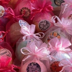 May include: A collection of bath bombs in various pastel colors, wrapped in sheer pink organza bags and tied with matching ribbons. Each bag is adorned with a round sticker that says "Thank You" in a decorative font.