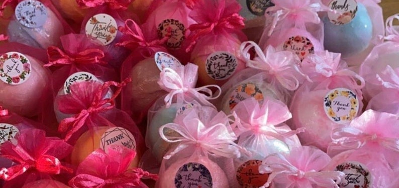 May include: A collection of bath bombs in various pastel colors, wrapped in sheer pink organza bags and tied with matching ribbons. Each bag is adorned with a round sticker that says "Thank You" in a decorative font.