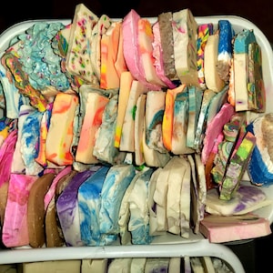 May include: A white plastic bin filled with colorful, handmade soap bars. The soaps are various shapes and sizes, with vibrant hues of pink, blue, orange, and green. Some bars have marbled patterns.
